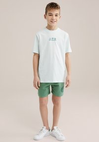 Svetlo mint zelena majica s tiskanim dizajnom, uparena s zelenim kratkim hlačama koje imaju detalj s logotipom. Bele patike upotpunjuju outfit.