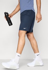 Αθλητικό σορτς Nike σε ναυτικό μπλε με λογότυπο σε λευκό, συνδυασμένο με λευκά αθλητικά παπούτσια, μαύρο παγούρι και αντίστοιχες λευκές κάλτσες.