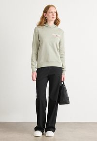Sudadera verde claro con un logo, combinada con pantalones negros y zapatillas blancas. Se sostiene un bolso acolchado negro en la mano derecha.