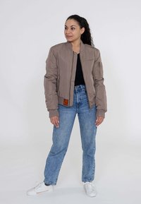 Giacca bomber marrone con zip frontale, polsini a costine e tasca laterale, abbinata a jeans blu in denim e sneakers bianche. Texture liscia.