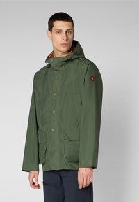 Vihreä vedenpitävä takki hupulla, napsulukolla, kaksi etutaskua ja oranssi vuori. Vasemmassa hihassa on logo.