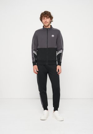 Mand med krøllet hår iført en sort og grå lynlåstræningsdragt med hvide striber og hvide sneakers, stående mod en ensfarvet hvid baggrund.