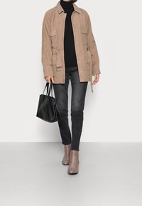 Veste en daim couleur tan avec poches, pull noir côtelé, jean gris foncé et bottines beige clair. Sac cabas noir tenu à la main.