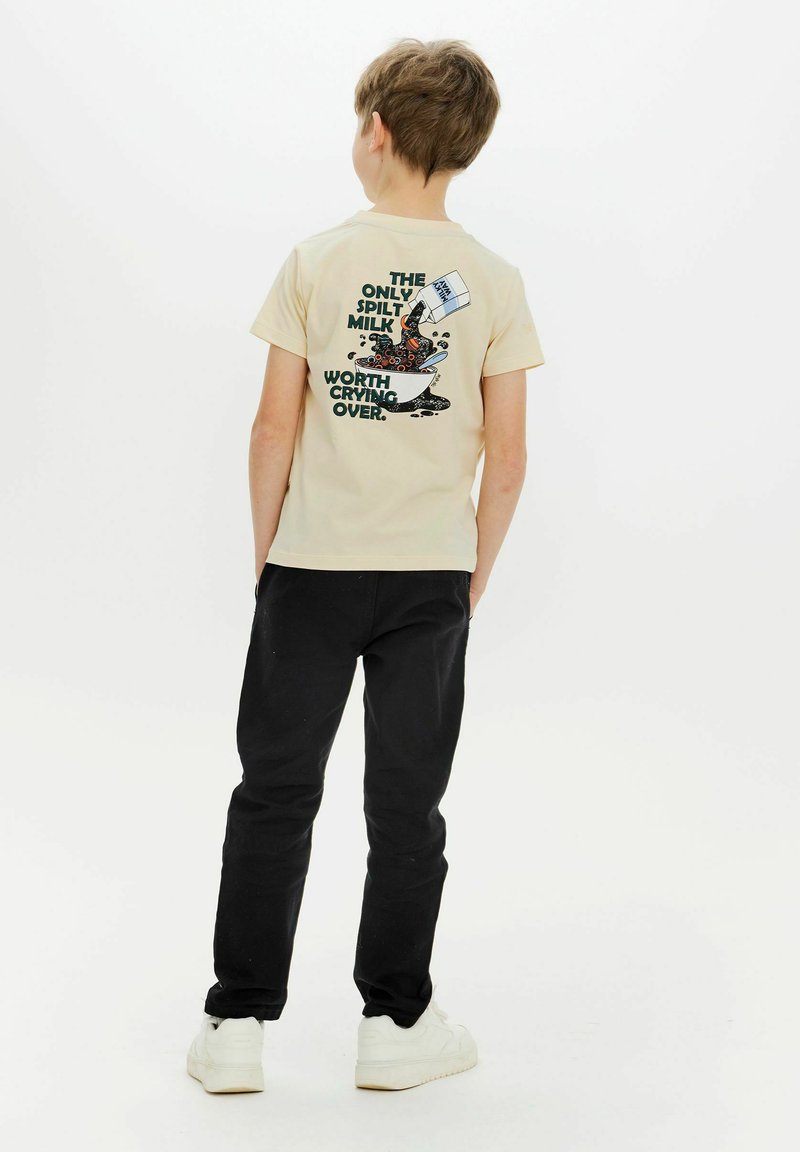 Camiseta de algodón beige de manga corta con un estampado gráfico en la parte trasera que presenta leche derramada y texto en azul oscuro y negro. Combinada con pantalones negros.