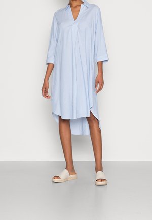 Woman wearing a light blue and white striped long-sleeve shirt dress with a high-low hem and beige open-toe slide sandals.