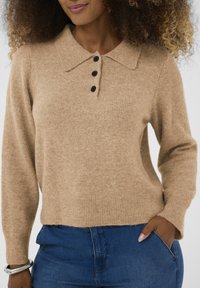 Bēša adīta džemperis ar apkakles kaklu, trim melniem pogām, ribotām piedurknēm un apakšmalu. Kombinēts ar ziliem džinsiem, kas piedāvā kabatu.