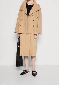 Beżowa dwurzędowa kurtka z dużym kołnierzem, w połączeniu z pasującą spódnicą do kolan. Czerwona torba na ramię i płaskie czarne sandały dopełniają stylizację.