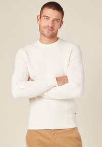 Weißer strukturierter Strickpullover mit Rundhalsausschnitt und gerippten Bündchen, kombiniert mit hellbraunen Cordhosen.