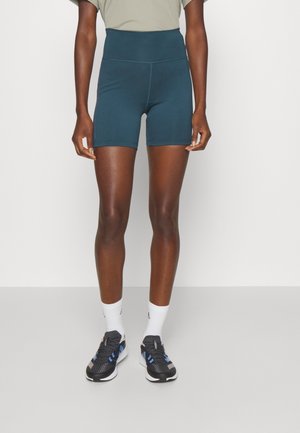 Persona que lleva pantalones cortos de ciclista de talle alto en color azul verdoso oscuro, calcetines blancos y zapatos deportivos negros, de pie sobre un fondo gris liso.