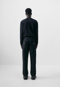 Chemise noire à manches longues et pantalon cargo, tous deux en tissu résistant, avec des poches et des accents dorés sur les manches, vus de dos.