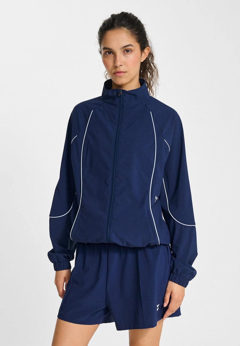 Marinblå idrottsjacka med full dragkedja, vita accentlinjer, hög krage och elastiska ärmslut, ihop med matchande shorts. Lättviktsmaterial.