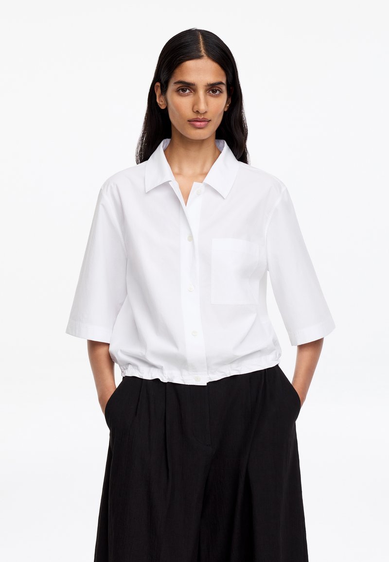 Femme aux longs cheveux noirs portant une chemise blanche à manches courtes boutonnée, rentrée dans un pantalon noir taille haute à jambes larges, les mains dans les poches.