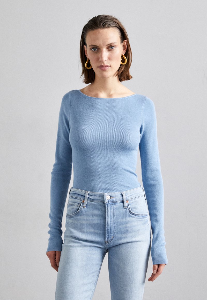 Vaaleansininen ribattu pitkähihainen paita, jossa venepääntie, yhdistettynä korkeavyötäröisiin vaaleansinisiin farkkuihin, joissa on hienovaraisia ompeluja ja napitusyksityiskohtia.