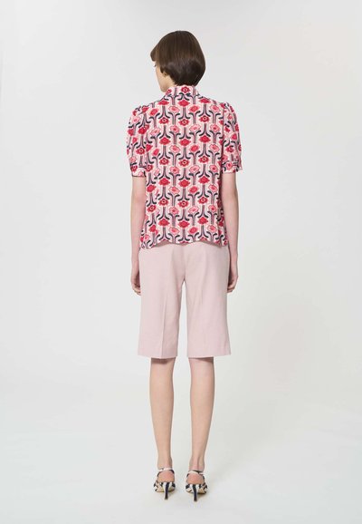 Femme debout de dos, portant un chemisier à manches courtes à motif floral, un short rose pâle longueur genou et des chaussures à talons avec un motif.