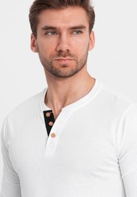 Mies, jolla on lyhyet ruskeat hiukset ja parta, pukeutunut valkoiseen pitkähiharaiseen henley-paitaan, jossa on musta nappilista, katsoo hieman syrjään.