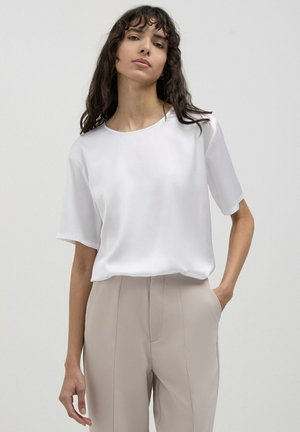 Blusa bianca di satin a maniche corte con scollatura rotonda, abbinata a pantaloni beige su misura, caratterizzati da una superficie liscia e una vestibilità morbida.
