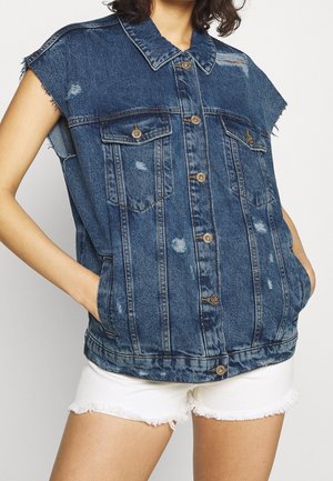 Nainen, jolla on päällään hihaton sininen kulutettu farkku-liivi, jossa on napitus, ja valkoiset rispaantuneet shortsit, kädet taskuissa.