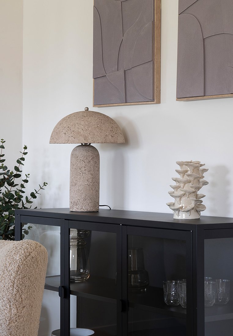 Braune Stehlampe mit runder Shade auf einem schwarzen Schrank. Weißes keramisches Dekorationsobjekt mit geschichtetem Design. Graue strukturierte Wandkunst darüber.