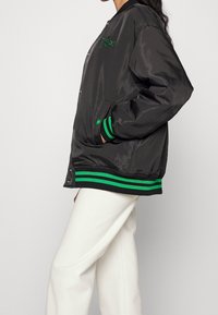 Svart bomberjacka med gröna randdetaljer, tryckknappar och broderad detalj. Tillverkad av glänsande tyg med ribbade ärmslut och nederkant.