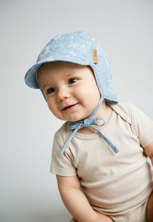 Smiling baby wearing a beige shirt and a blue hat with white constellations, tied under the chin, against a light background.