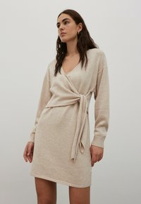Robe en tricot beige avec un design enveloppant, décolleté en V profond, manches longues et un lien à la taille ; texture douce et poignets côtelés.