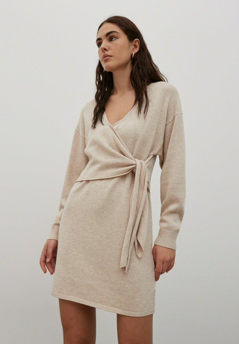 Robe en tricot beige avec un design enveloppant, décolleté en V profond, manches longues et un lien à la taille ; texture douce et poignets côtelés.