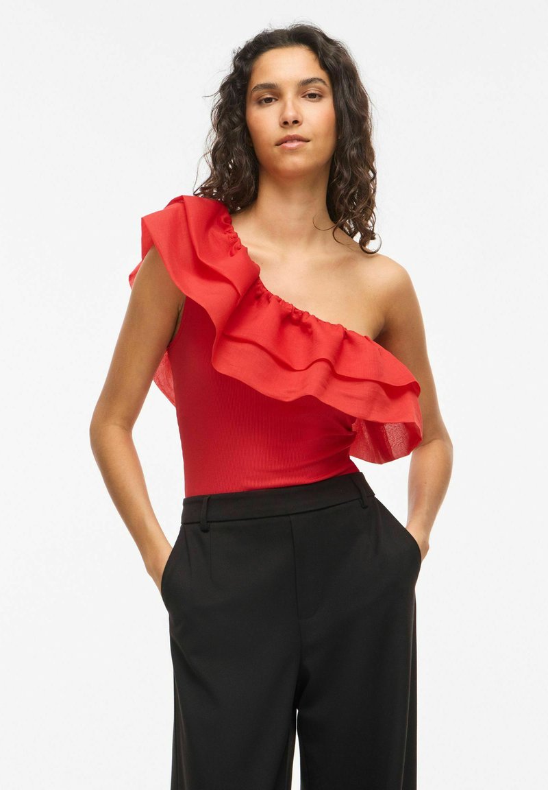 Femme aux cheveux bouclés portant un haut rouge sans manches à volants et un pantalon noir taille haute, mains dans les poches, sur fond blanc uni.
