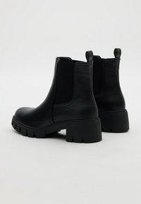 Botas de cuero negras hasta el tobillo con suelas gruesas, paneles laterales elásticos y tiradores en la parte posterior sobre un fondo blanco liso.