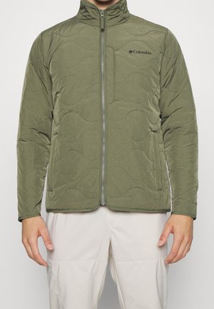 Mand iført en olivengrøn quiltet Columbia-jakke med lynlås foran og lyse beige bukser foran en ensfarvet baggrund.