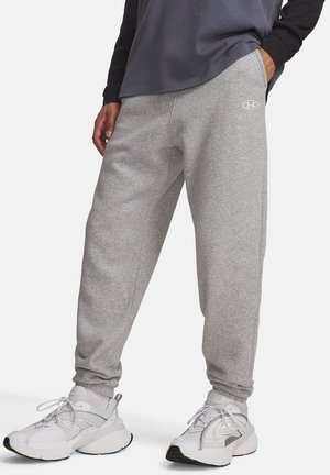 Person iført lysegrå joggingbukser med logo, hvide sneakers, sorte langærmede, og en mørkegrå trøje, stående med den ene hånd i lommen.