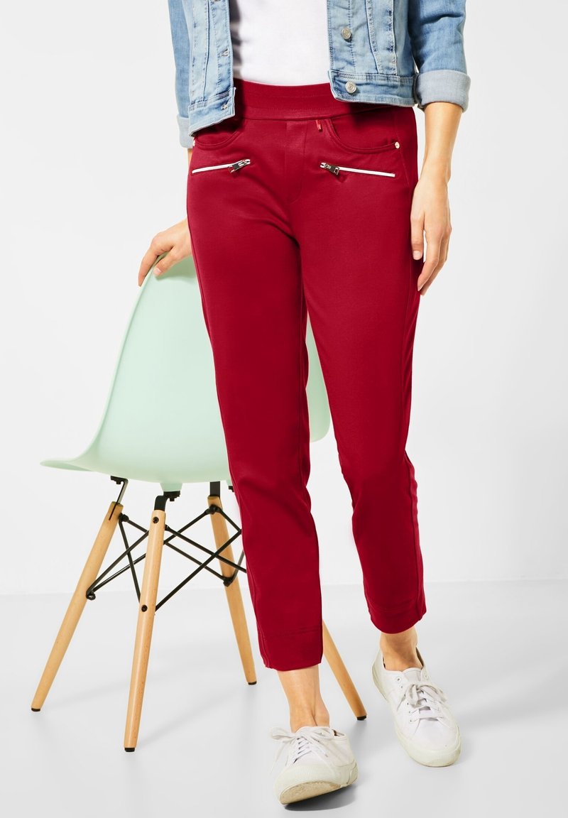 Rode skinny jeans met een zachte textuur en elastische tailleband, voorzien van twee ritszakken aan de voorkant. Gepaard met witte sneakers.