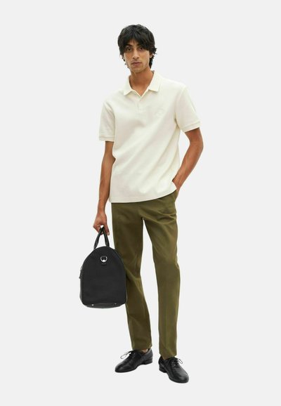 Jonge man in een witte korte mouwen polo, olijfgroene broek, zwarte nette schoenen, houdend een zwarte handtas, staande tegen een effen achtergrond.