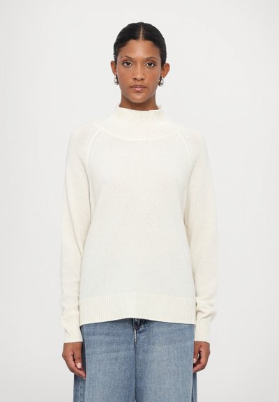 Pull en col roulé couleur crème avec des poignets et un ourlet côtelés, présentant une texture douce et une coupe légèrement décontractée. Porté avec un jean en denim bleu clair.