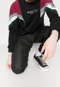 Persona de rodillas vistiendo una sudadera negra, burdeos, verde y gris, pantalones negros, calcetines blancos y zapatillas negras con cordones blancos.