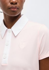 Κοντινό πλάνο ενός ατόμου που φοράει ροζ πουκάμισο πόλο με λευκό γιακά και κουμπιά, με ένα μικρό κεντητό λογότυπο στο στήθος.