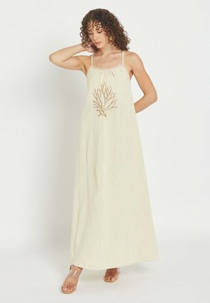 Mujer con cabello rizado vistiendo un vestido maxi beige sin mangas con tirantes finos y bordado de ramas de coral en el pecho, de pie sobre fondo blanco.