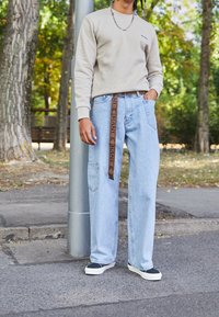 Sudadera gris claro, jeans de mezclilla azul holgados con un cinturón de logo marrón y zapatos negros sin cordones en una acera cerca de un poste de calle.