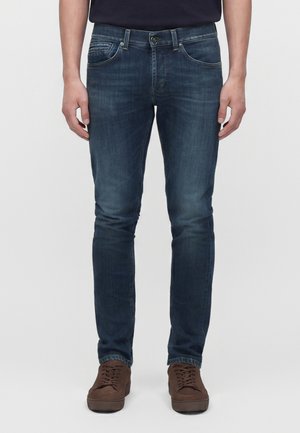 Άντρας που φοράει σκούρα μπλε slim-fit τζιν και καφέ casual παπούτσια με κορδόνια, στέκεται μπροστά σε απλό ανοιχτό φόντο.