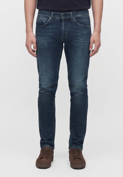 Άντρας που φοράει σκούρα μπλε slim-fit τζιν και καφέ casual παπούτσια με κορδόνια, στέκεται μπροστά σε απλό ανοιχτό φόντο.