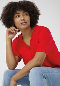 Blusa rossa con scollatura arricciata e maniche corte. Il materiale sembra liscio; la modella è seduta, indossando jeans azzurri.