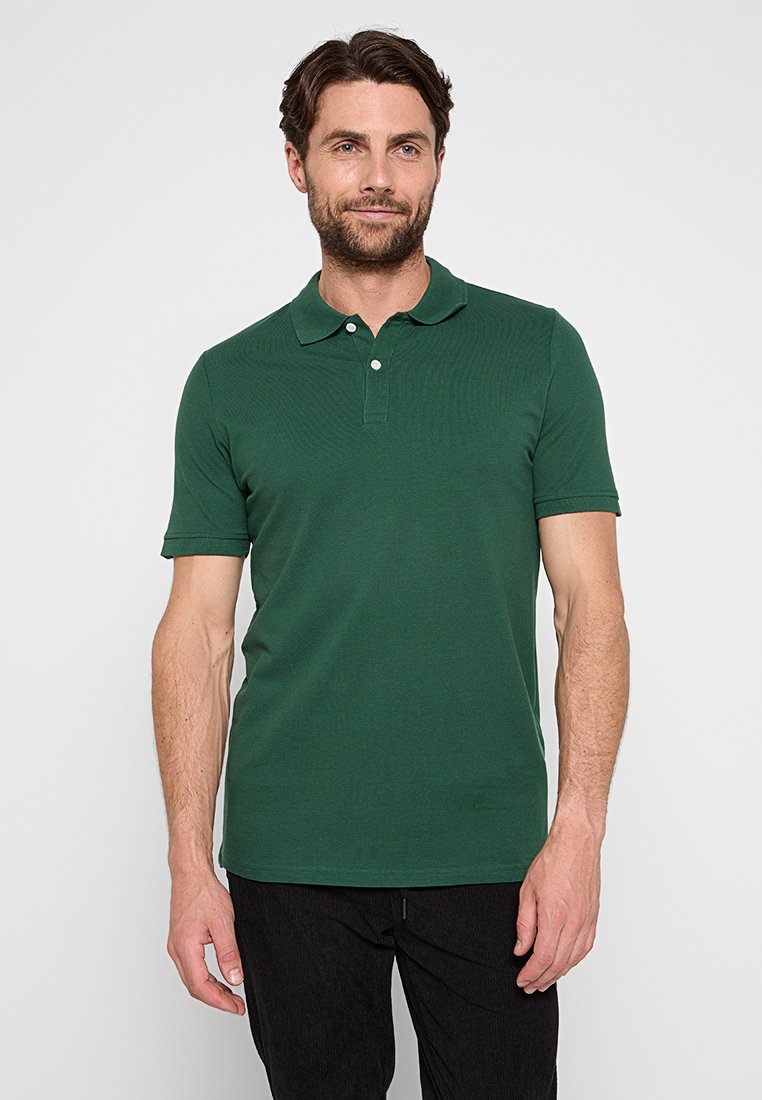 Polo shirt court vert foncé avec un col, une patte de boutonnage à deux boutons et un tissu texturé. Porté avec un pantalon foncé.