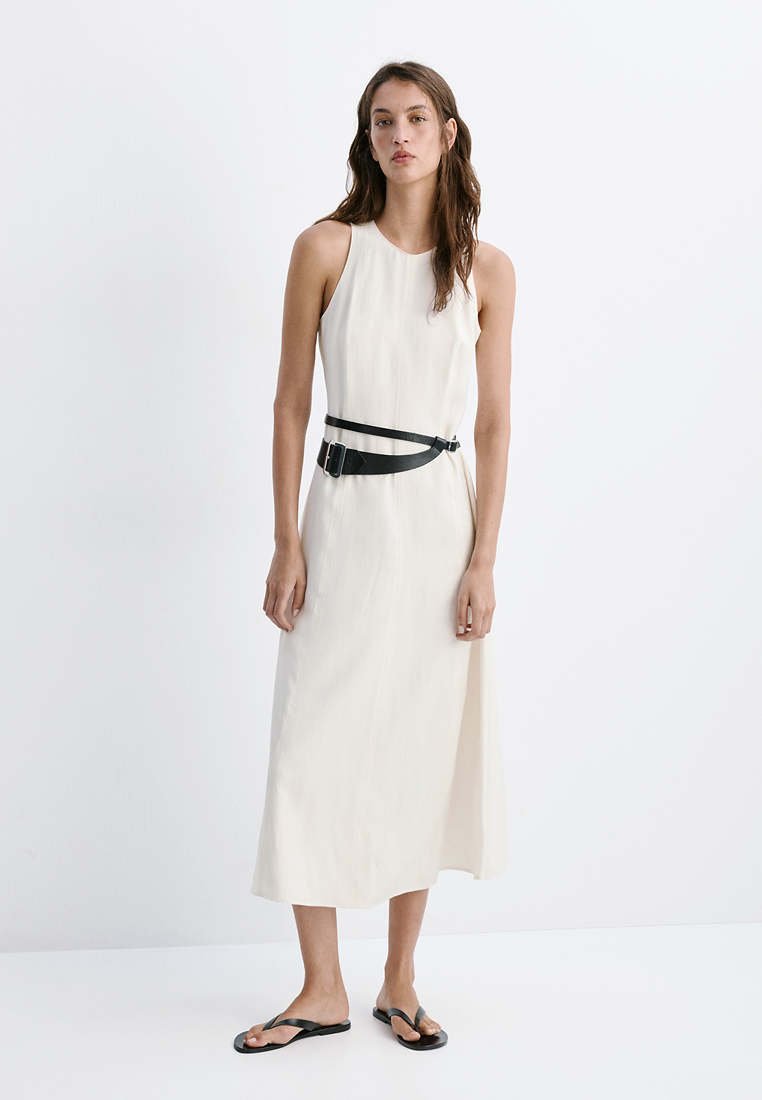 Femme aux longs cheveux bruns porte une robe midi blanche sans manches avec ceinture noire et sandales plates noires, debout devant un fond blanc uni.