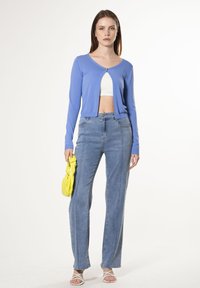 Cárdigan de manga larga azul claro con un cierre frontal, combinado con unos jeans de mezclilla de pierna recta y cintura alta y sandalias blancas de tiras. Lleva un bolso amarillo.
