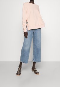 Lichtroze sweatshirt met zijruffels, gecombineerd met blauwe wijde denim jeans en beige sandalen met bandjes. Eenvoudig, casual outfitontwerp.