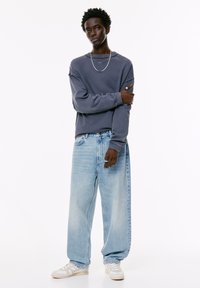 Jonge man met kort krullend haar, gekleed in een losse donkergrijze sweatshirt, wijde lichtblauwe spijkerbroek, witte sneakers en zilveren accessoires.