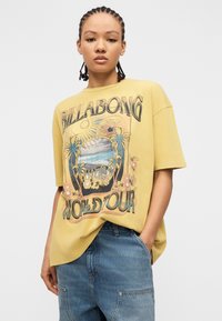 Νεαρός με πλεξούδες, που φορεί κίτρινο μπλουζάκι Billabong World Tour 1973 με γραφικά και μπλε τζιν, με το ένα χέρι στην τσέπη.