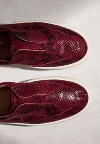 Paire de chaussures slip-on bordeaux avec un motif en cuir de serpent texturé et brillant et des semelles en caoutchouc blanc sur une surface gris clair.