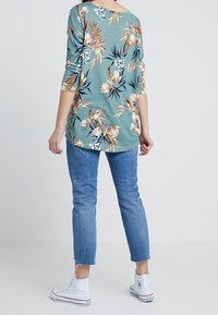 Bluzka w zielono-niebieskim wzorze kwiatowym o luźnym kroju i szerokim dekolcie, zestawiona z krótkimi niebieskimi jeansami i białymi trampkami na wysokiej podeszwie.