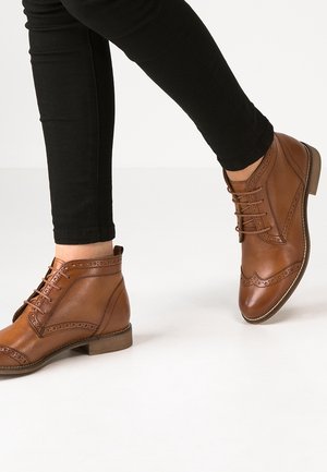 Bottines en cuir marron avec détails brogues portées avec un pantalon noir ajusté sur fond blanc.