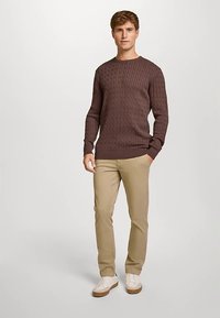 Maglione marrone lavorato a maglia con motivo a rilievo, scollo rotondo, abbinato a pantaloni beige e sneakers bianche. La modella è in piedi, eretta.